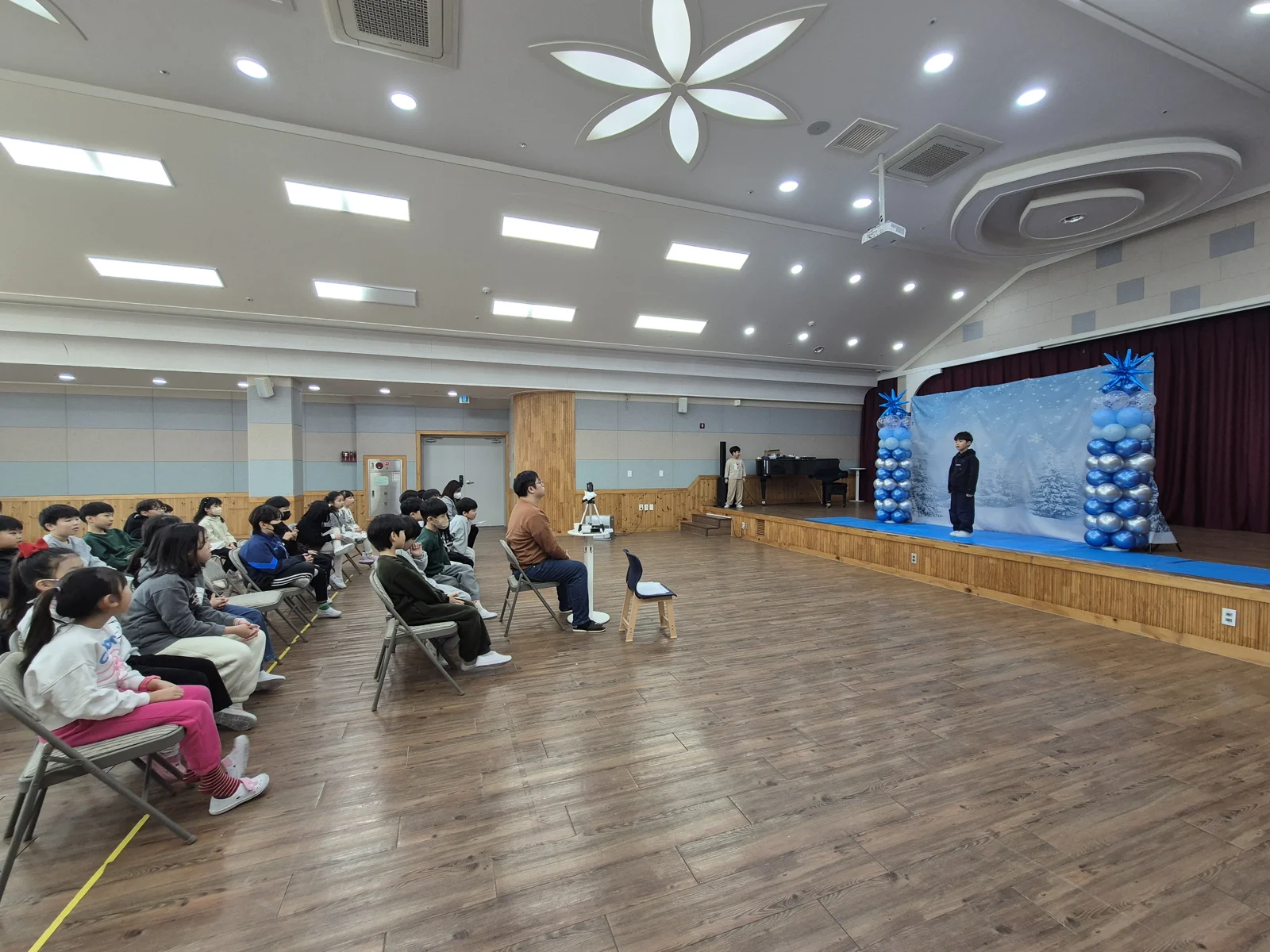 강당 무대에서 발표하는 아이와 지켜보는 친구들
